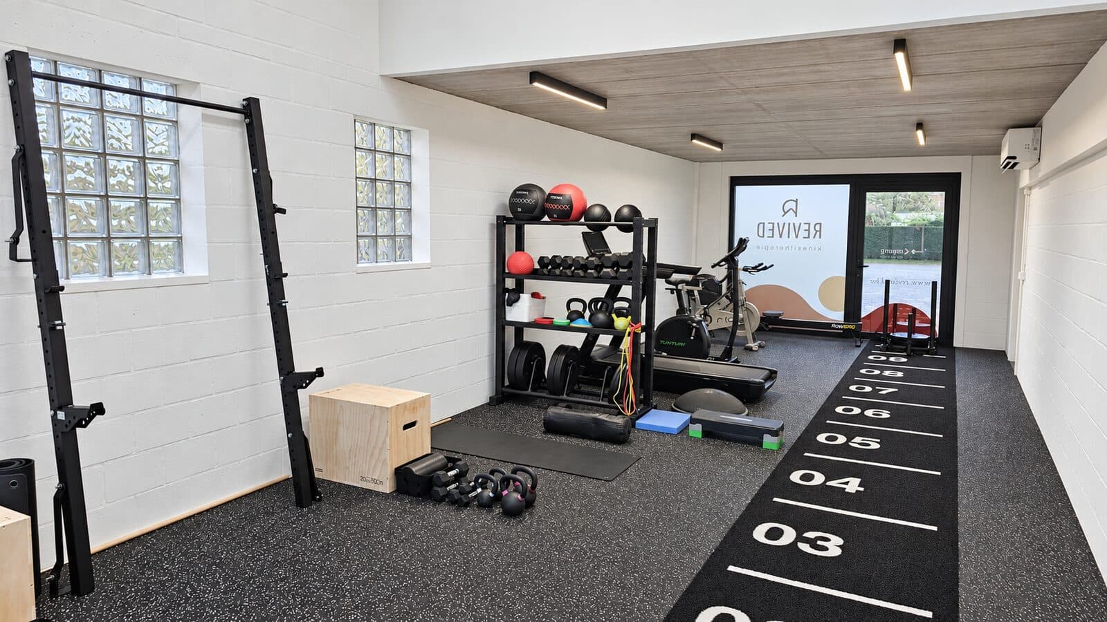 Overzicht van de oefenzaal met fitnessmateriaal en de Revived-ramen